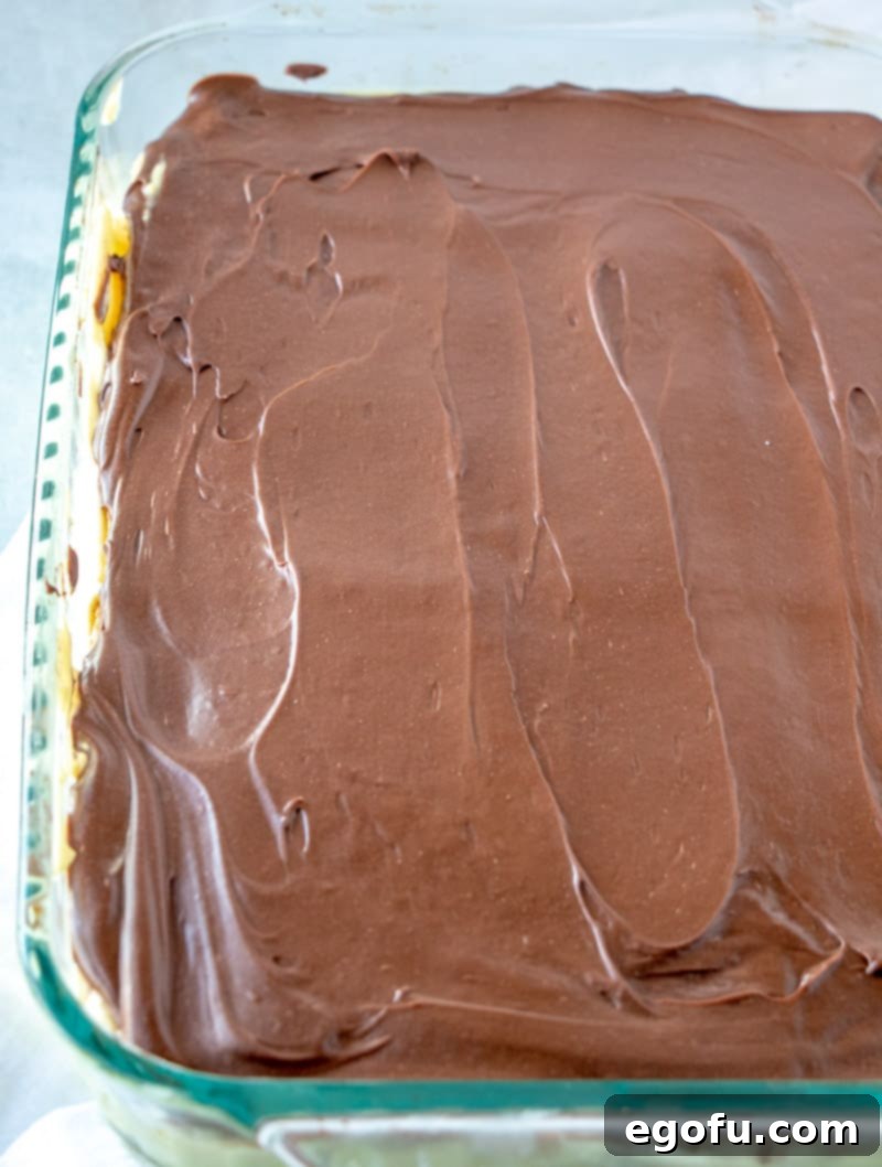 Smooth chocolate frosting being evenly spread across the top layer of graham crackers on the Eclair Cake in a baking dish.