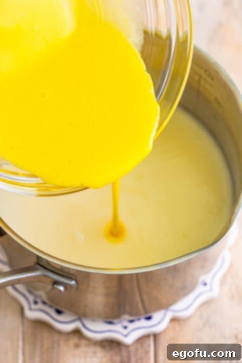 Golden Banana Dream Pie 21 The tempered egg yolk mixture being gently poured into the simmering pudding base in a saucepan, ensuring a smooth custard.