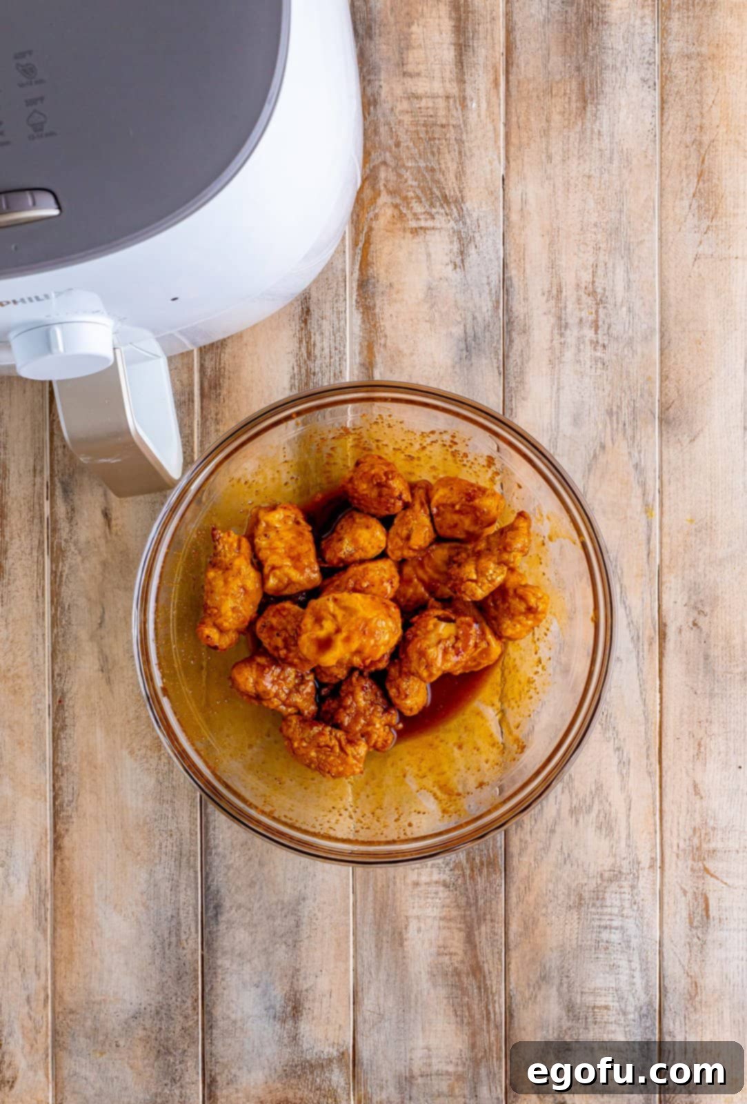 Cooked chicken nuggets being tossed in homemade Nashville hot sauce in a clear mixing bowl, ensuring even coating.