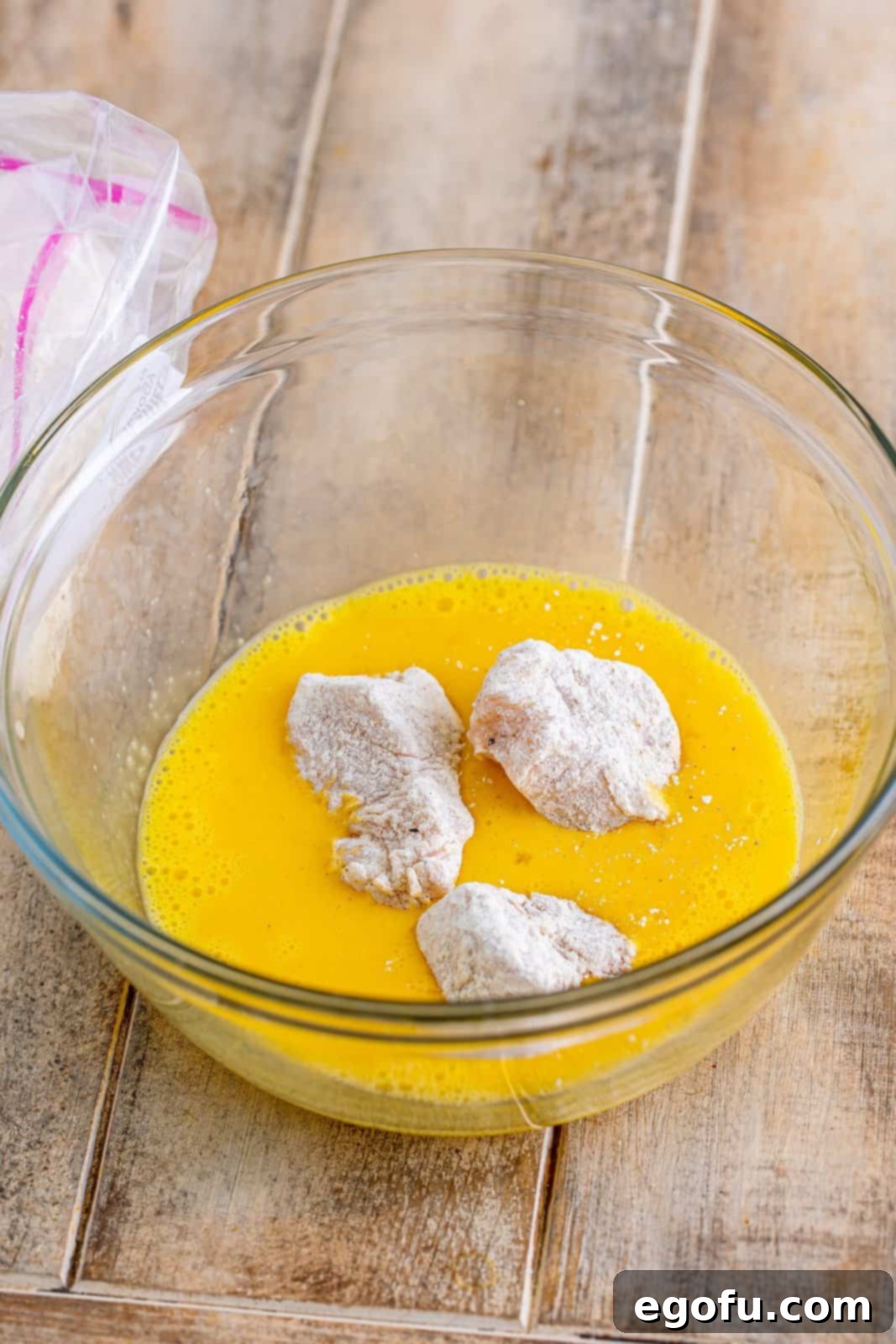 Flour-coated chicken pieces being dipped into whisked egg mixture in a clear bowl for the second step of the dredging process.