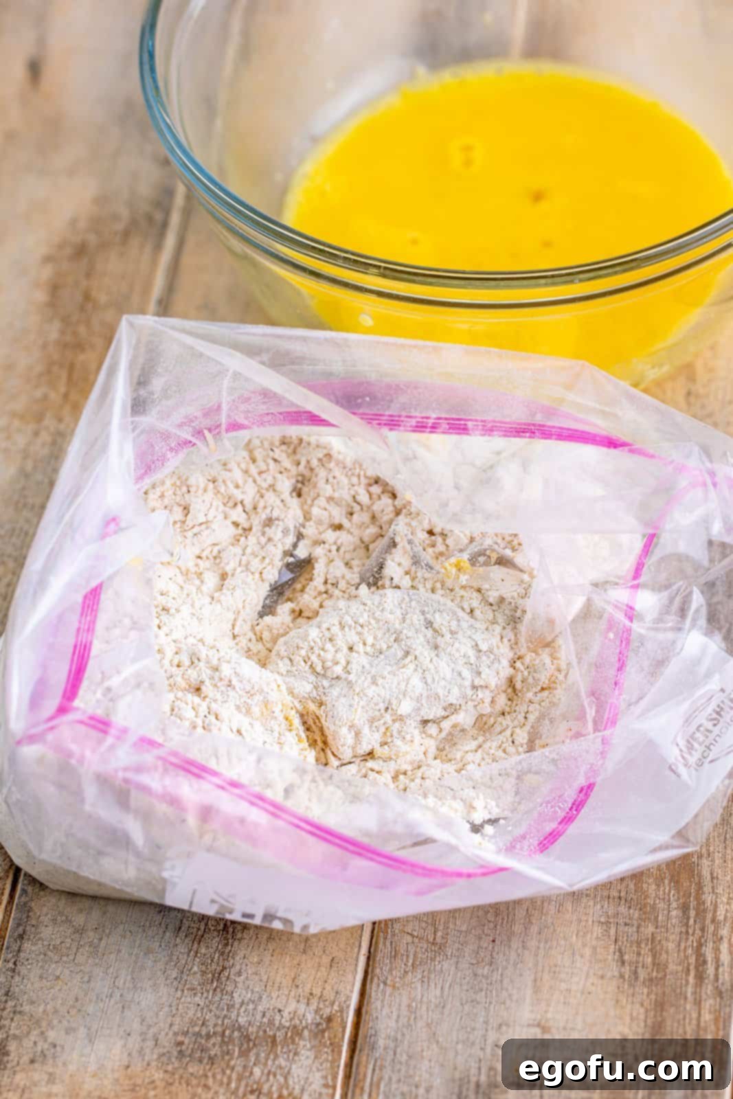 Egg-coated chicken breast returning to the flour mixture in a plastic bag for a second coating, ensuring a thick, crispy crust.