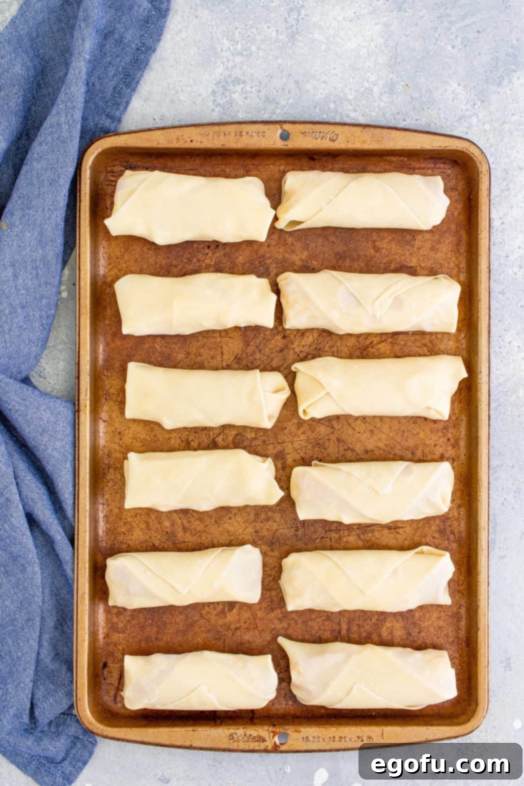 A collection of perfectly rolled egg rolls neatly arranged on a baking pan, covered with a clean kitchen towel to keep them fresh.
