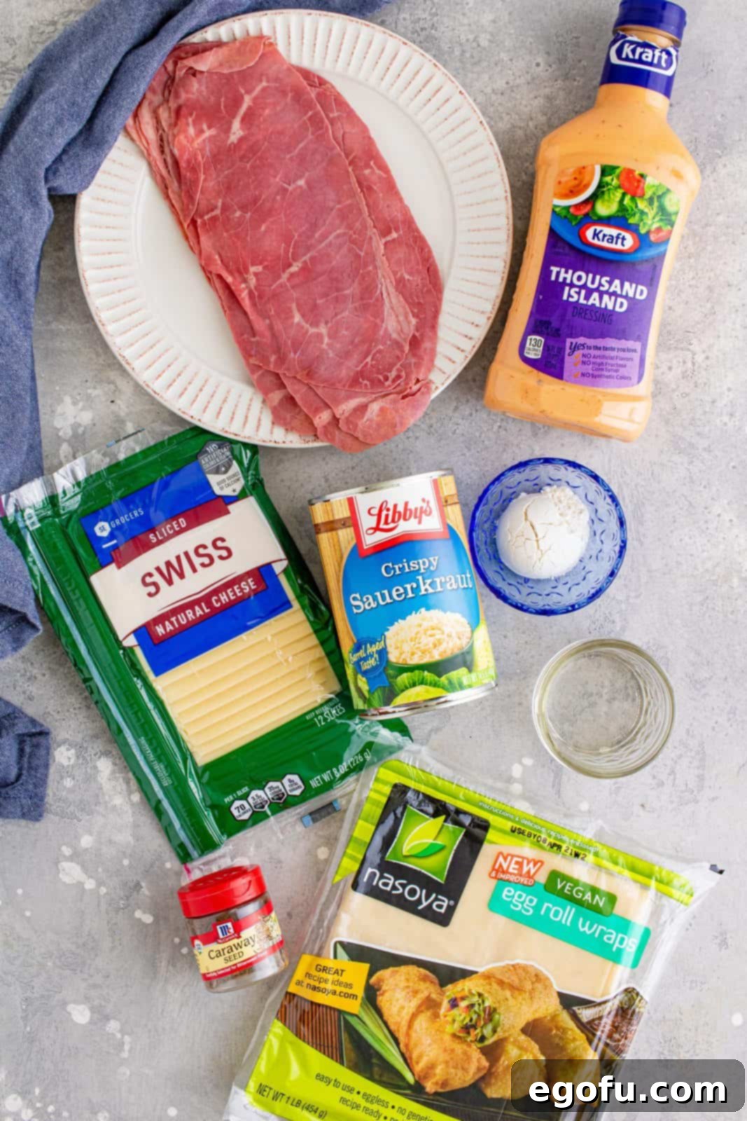 Ingredients laid out: a bowl of sauerkraut, Thousand Island dressing, caraway seeds, a pile of all-purpose flour, a glass of water, a stack of egg roll wrappers, slices of swiss cheese, a mound of deli corned beef, a bottle of oil, and fresh scallions for garnish.
