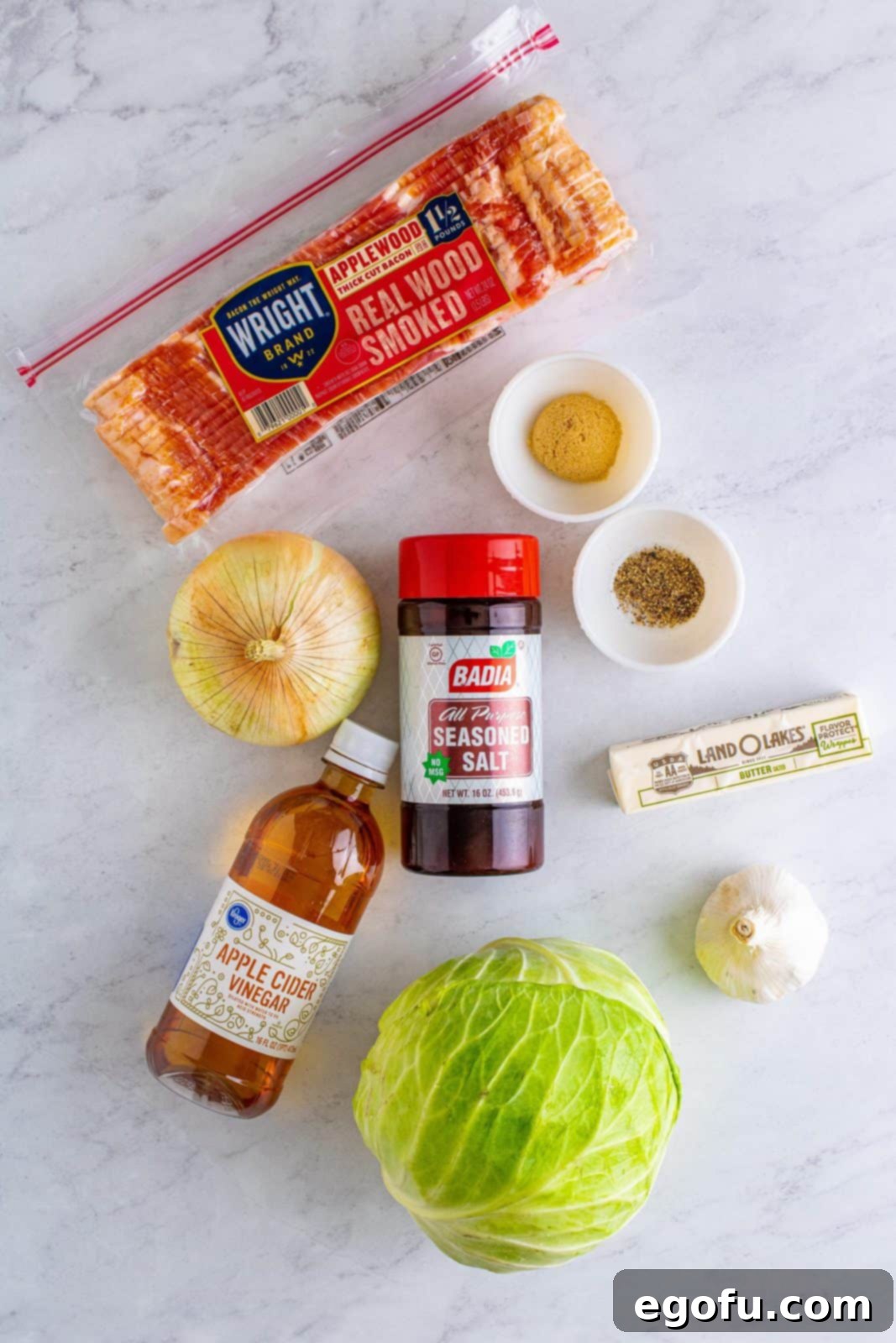 Crispy Bacon and Caramelized Cabbage 4 A flat lay photograph showcasing all the fresh ingredients needed for Southern Bacon Fried Cabbage: thick-cut bacon, a whole onion, garlic cloves, a head of green cabbage, a bottle of apple cider vinegar, a bowl of light brown sugar, seasoned salt, black pepper, and a stick of salted butter.