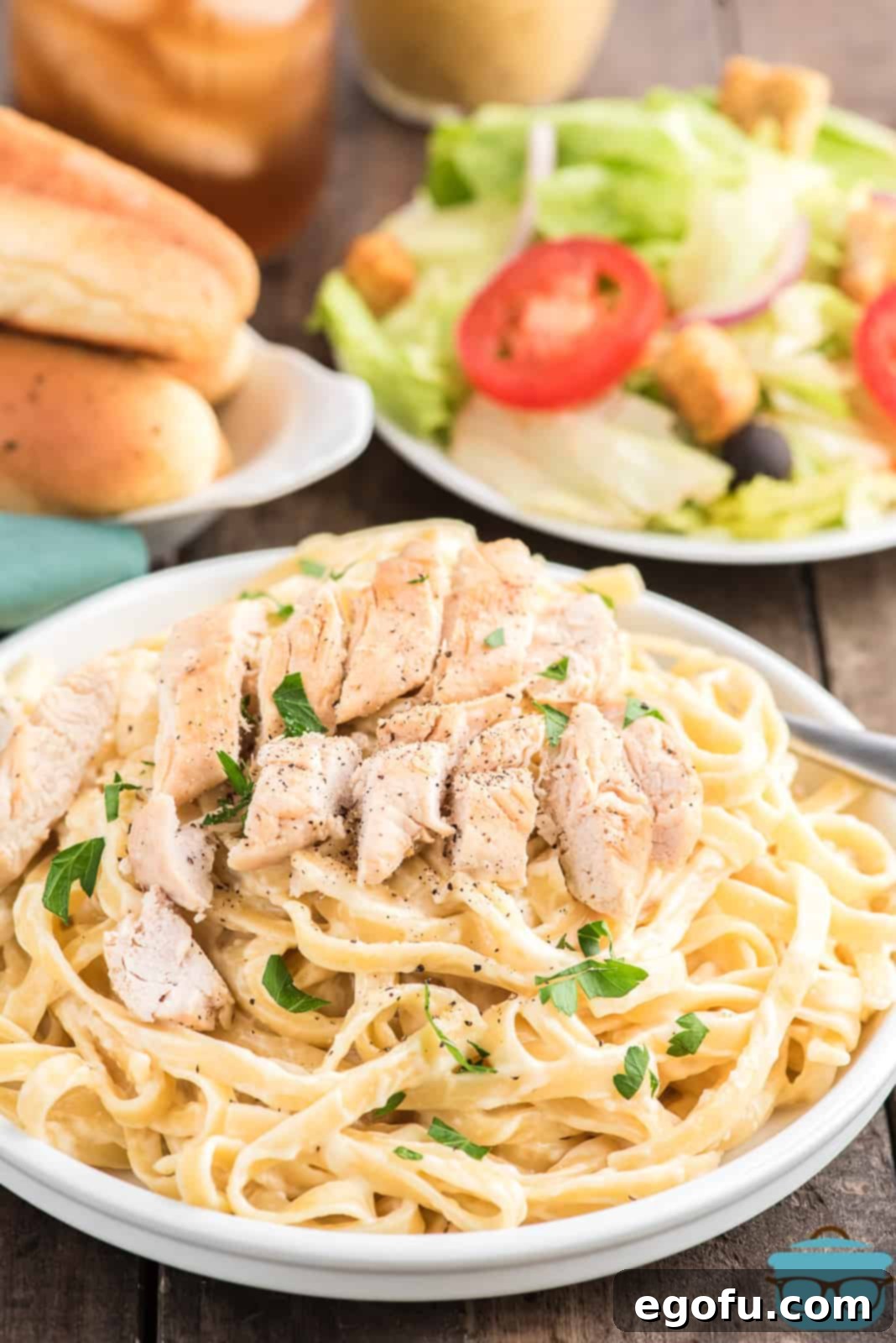 Finished Copycat Olive Garden Chicken Alfredo in a bowl, garnished with parsley, with a side salad and breadsticks in the background.