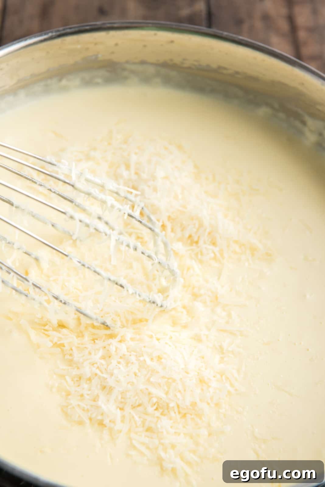 Shredded Parmesan and Romano cheeses being stirred into the simmering cream sauce in a skillet.