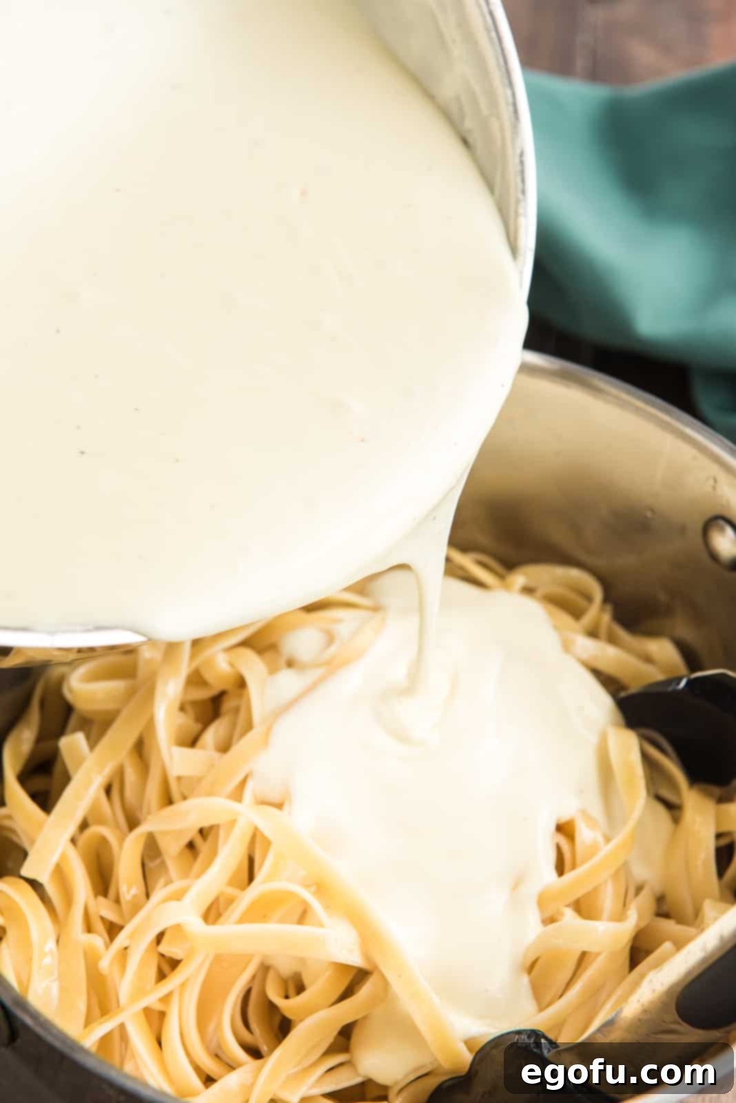 Creamy Alfredo sauce being poured over a mound of drained fettuccine pasta in a large pot.