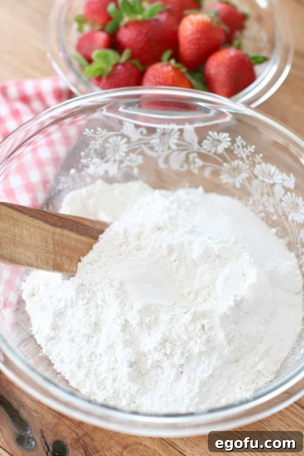 A clear glass bowl filled with the dry biscuit ingredients: baking powder, granulated sugar, and all-purpose flour, awaiting further mixing, with a wooden spoon resting nearby.