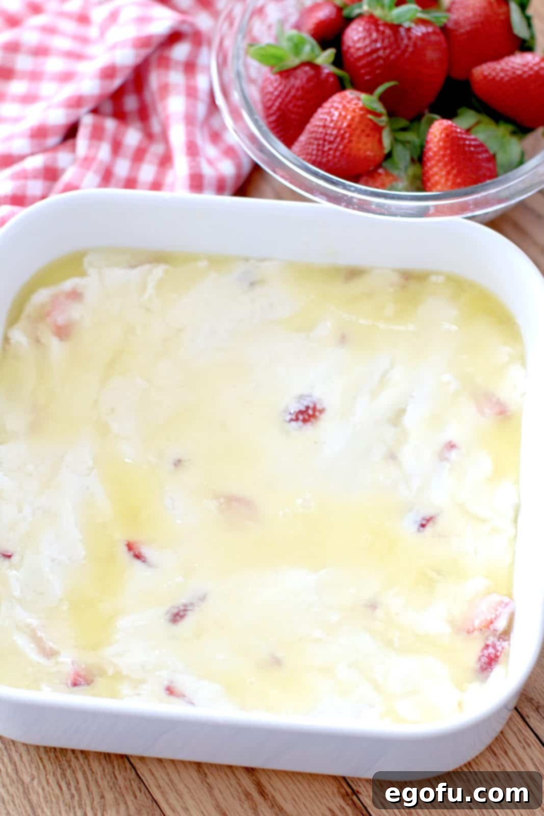 The soft biscuit dough, studded with strawberries, spread evenly into a white square baking dish, resting atop a pool of melted butter, ready for its transformation.